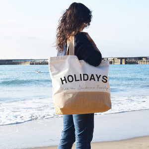 Person holding a tote bag with 'HOLIDAYS' text on a beach