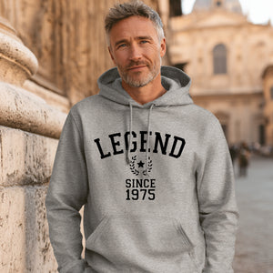 Man wearing a gray hoodie with 'LEGEND SINCE 1975' text, standing in front of stone architecture.